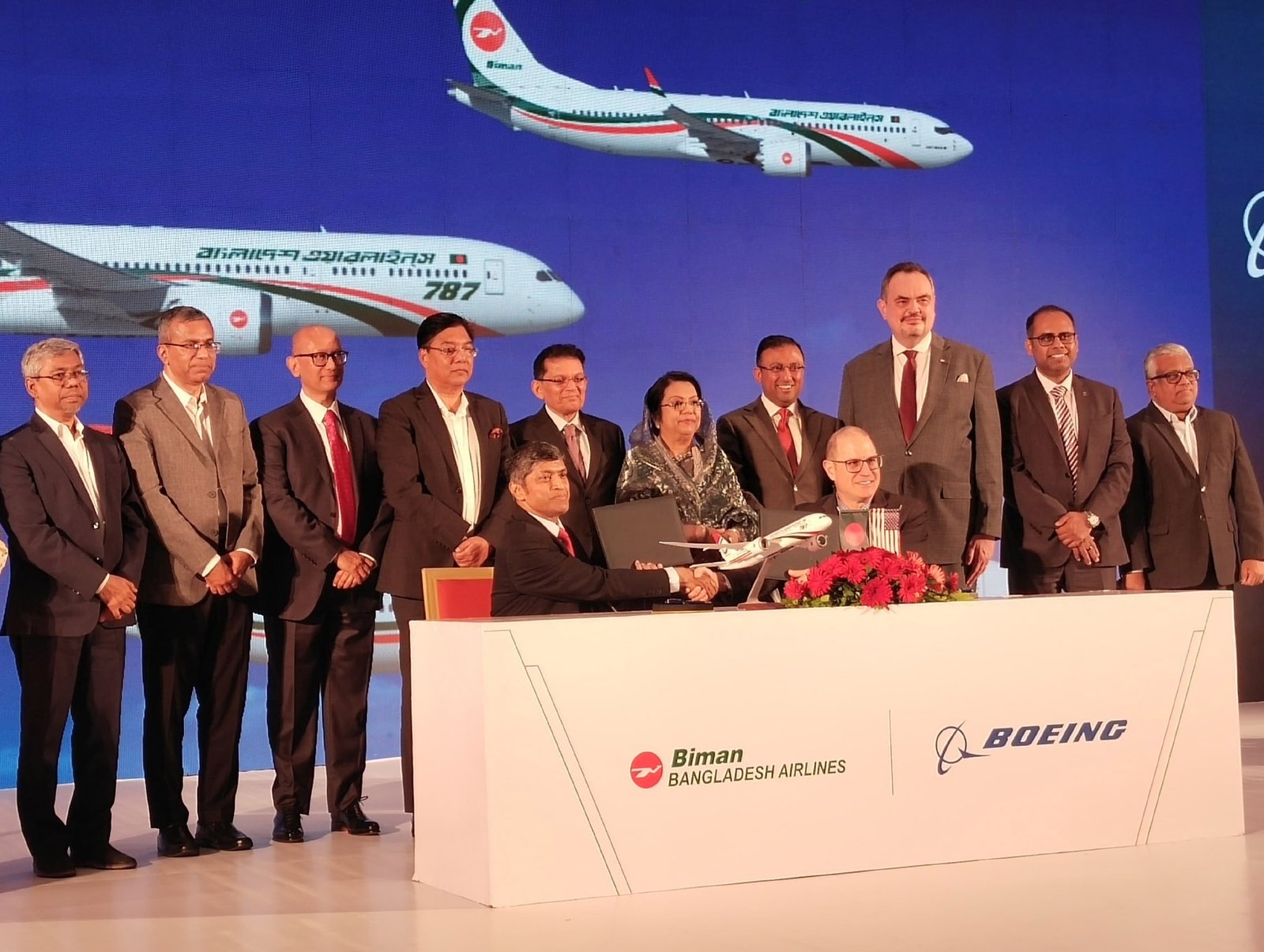 Officials signing an aircraft acquisition agreement at a formal ceremony with aircraft imagery behind them.